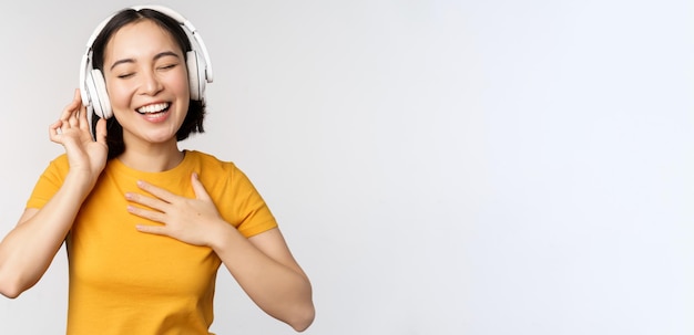Chica asiática feliz bailando escuchando música en auriculares y sonriendo de pie en camiseta amarilla contra...