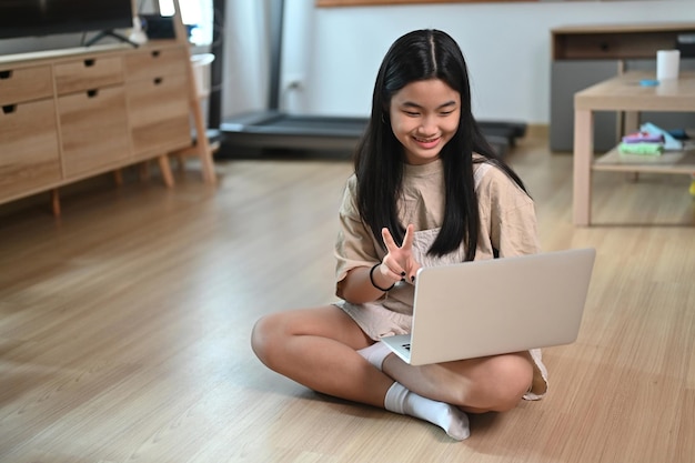 Chica asiática feliz que tiene una videollamada con una computadora portátil mientras está sentada en el piso de la sala de estar.