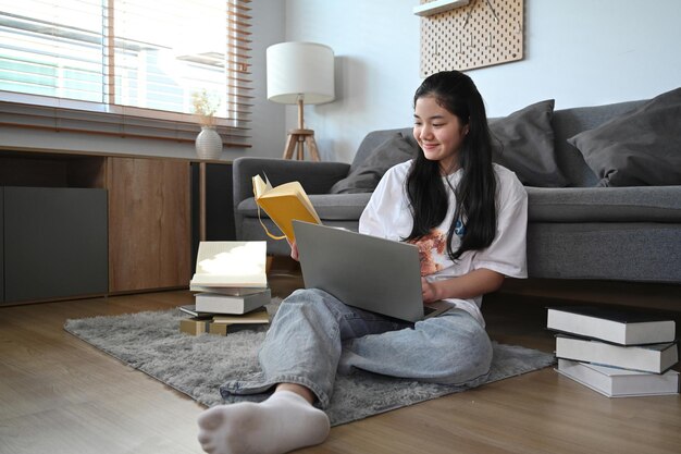 Chica asiática feliz sentada en la sala de estar y aprendiendo en línea en la computadora portátil