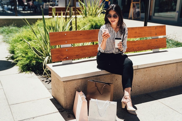 Chica asiática feliz usando un teléfono inteligente en el centro comercial de la ciudad sentado en un banco en stanford. mujer joven que usa un teléfono celular sosteniendo un café chateando un mensaje de texto en línea con bolsas de compras en el suelo descansando después de la tienda.