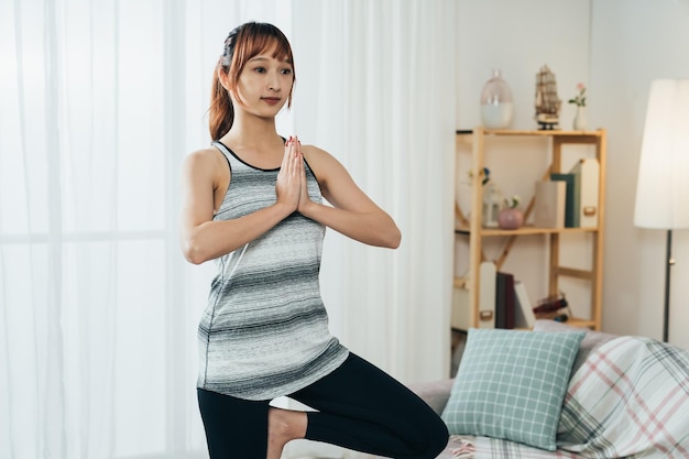 Una chica asiática en forma de retrato en ropa deportiva está practicando la pose del árbol de yoga durante el entrenamiento matutino en la sala de estar de casa.