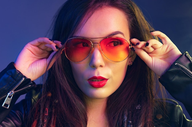 Chica asiática con gafas de sol y chaqueta de cuero negra posando contra un fondo azul oscuro
