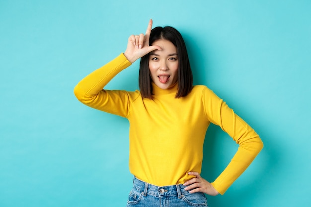 Chica asiática ganadora burlándose del equipo perdido, mostrando el signo de perdedor en la frente y lengua ignorante, de pie sobre fondo azul.