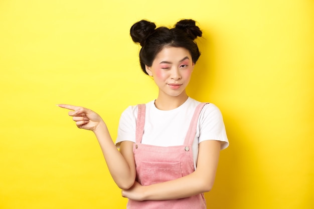 Chica asiática glamorosa con maquillaje brillante, apuntando con el dedo hacia la izquierda y guiñando un ojo a la cámara, insinuación de un buen trato promocional, de pie en amarillo