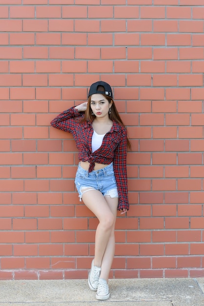 Chica asiática hipsters en pared de ladrillo naranjaestilo de vida de adolescente modernoFeliz dama posando para tomar una fotogente de Tailandia vistiendo camisas a cuadros y pantalones cortos de jeans