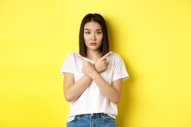 Chica asiática indecisa necesita ayuda con la elección, apuntando con el dedo hacia los lados y luciendo confundida, parada sobre fondo amarillo.