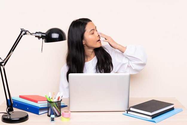 Chica asiática en un lugar de trabajo con una computadora portátil en la pared beige bostezando y cubriendo la boca abierta con la mano