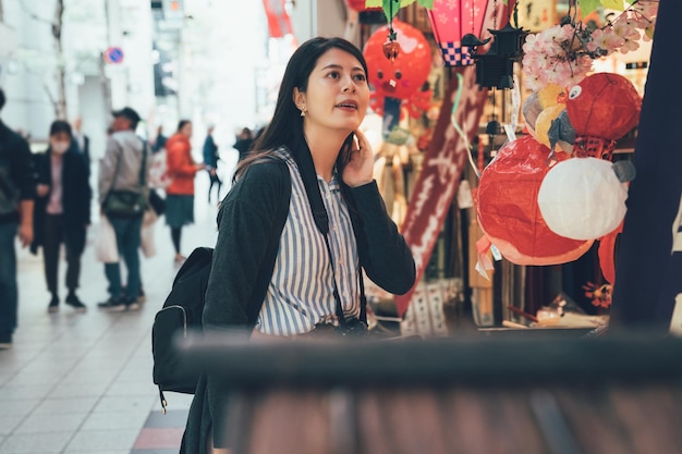 chica asiática mirando de compras en un vendedor ambulante en kuromon ichiba en osaka, japón. joven china comprando en el mercado tradicional local japonés en un puesto de farolillos de juguete. fotógrafo de viajes de visita.