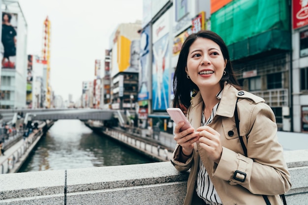 Chica asiática moderna de la ciudad urbana que usa la aplicación de teléfono sonriendo caminando en el puente cruza el canal en dotonbori osaka japón. Concepto de estilo de vida de viaje de negocios para adultos jóvenes felices en primavera con informal elegante.