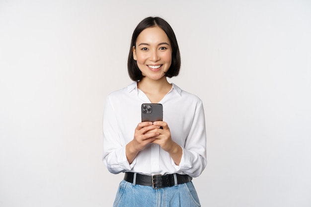 Chica asiática moderna y elegante usando una aplicación de teléfono móvil chateando por teléfono celular y sonriendo de pie en una blusa blanca contra el fondo del estudio