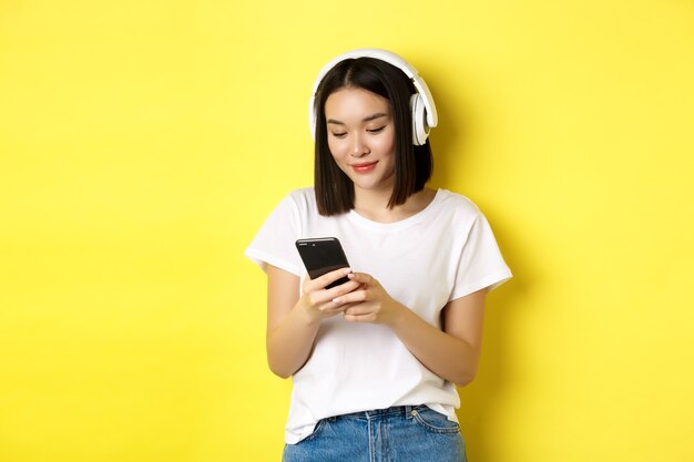 Chica asiática moderna escuchando música en auriculares inalámbricos, leyendo la pantalla del teléfono inteligente y sonriendo, de pie en camiseta blanca sobre fondo amarillo