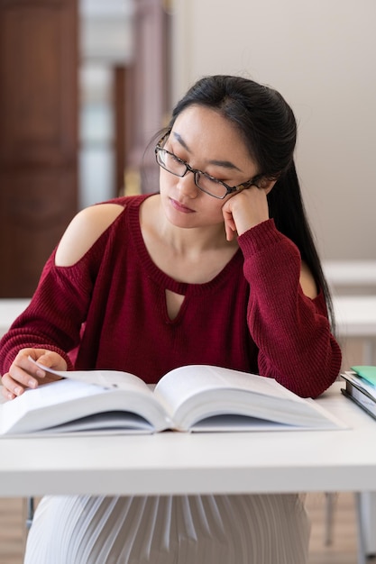 Chica asiática nerd leyendo libros de texto mientras se prepara para el examen o prueba en la sala de lectura de la biblioteca