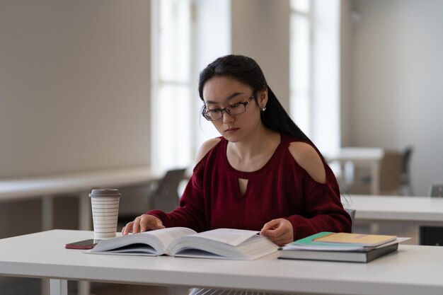 Chica asiática nerd leyendo libros de texto mientras se prepara para el examen o prueba en la sala de lectura de la biblioteca