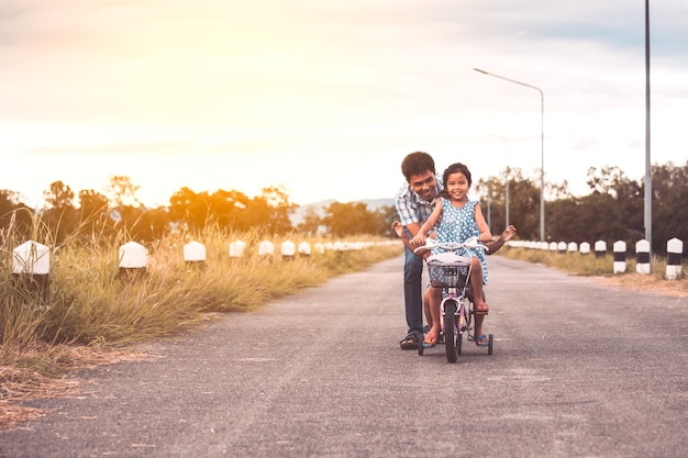 Chica asiática niño divirtiéndose para andar en bicicleta con el padre ayuda a empujarlos juntos