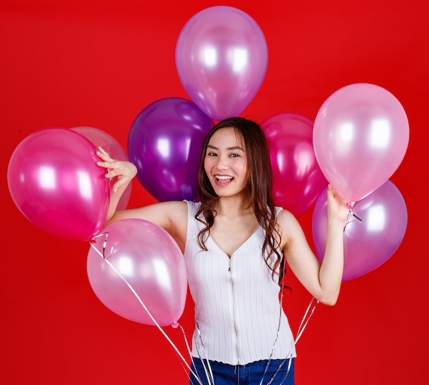 Chica asiática de pelo largo oscuro lindo y hermoso sosteniendo y jugando con globos de aire de colores con una sonrisa divertida y feliz sobre fondo rojo, tiro de luz de estudio.