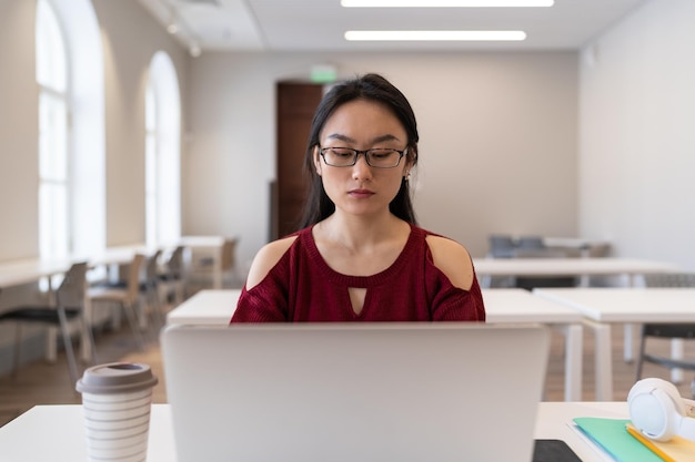 Chica asiática pensativa enfocada en gafas estudiando en línea en una computadora portátil mientras se sienta en la mesa de la biblioteca