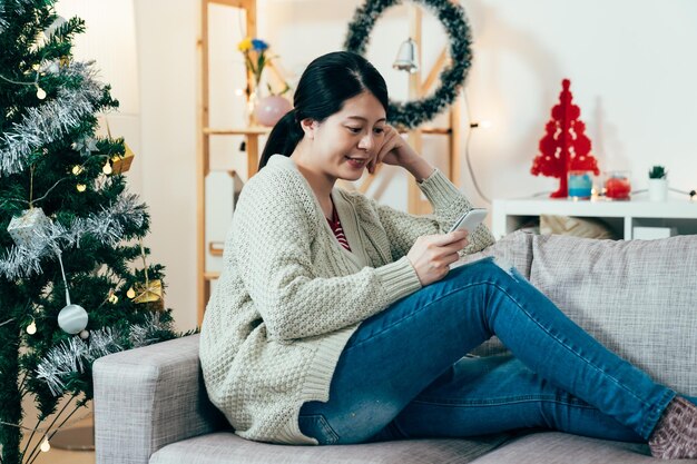 una chica asiática retratada sentada en un sofá sosteniendo un móvil está viendo videos en línea en un estado de ánimo relajante. tecnología y concepto de estilo de vida genuino