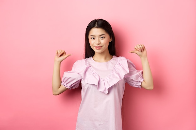 Chica asiática segura sonriendo, señalando a sí misma, autopromoción de logros personales, de pie en vestido sobre fondo rosa.