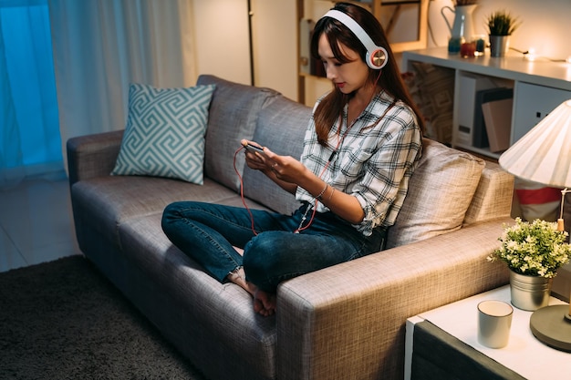 una chica asiática sentada en un sofá con ropa informal está sosteniendo y viendo videos en línea en su smartphone con auriculares durante la noche en una acogedora sala de estar en casa