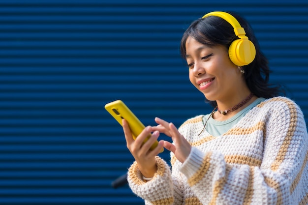 Chica asiática sonriendo escuchando música con auriculares amarillos en un fondo universitario azul