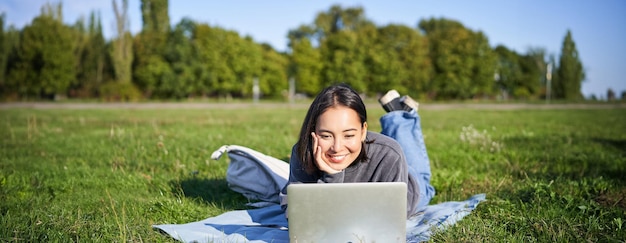 Una chica asiática sonriente acostada en el parque en la hierba viendo una película o video en una computadora portátil mirando la pantalla con