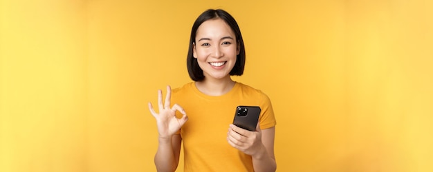 Chica asiática sonriente feliz sosteniendo el teléfono móvil y mostrando bien recomendando la aplicación en el teléfono inteligente