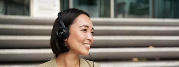 Chica asiática sonriente riendo escuchando música con auriculares sentado al aire libre estudiante universitario disfrutando gratis