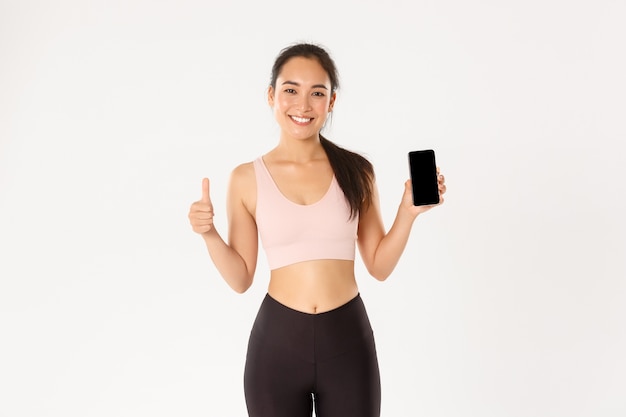 Chica asiática sonriente satisfecha de fitness, deportista atractiva que muestra el pulgar hacia arriba y la pantalla del teléfono inteligente, recomienda la aplicación de seguimiento de entrenamiento móvil.