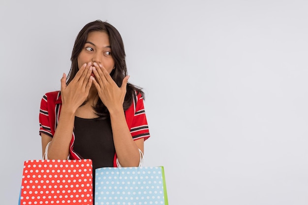 Chica asiática sorprendida cerrando la boca con las manos mientras trae las bolsas de compras en backgr aislado