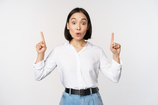 Chica asiática sorprendida señalando con el dedo hacia arriba mostrando información de banner superior o publicidad de pie sobre fondo blanco en blusa