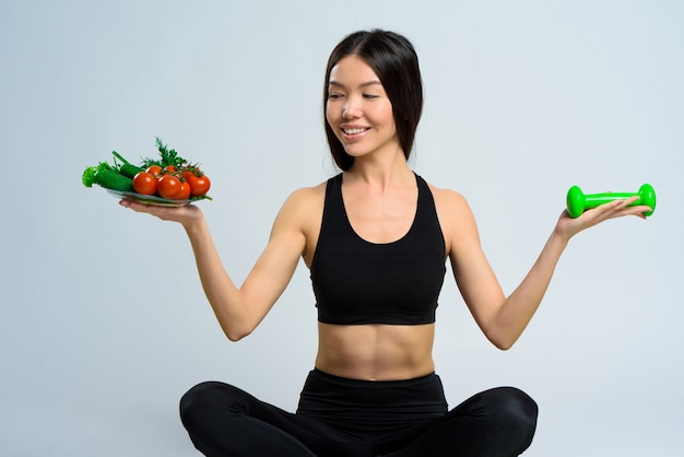 Chica asiática tiene pesas y plato de verduras.