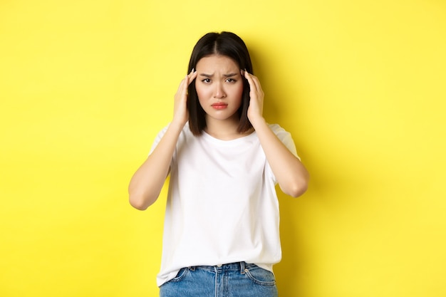 Chica asiática triste tocando la cabeza y frunciendo el ceño, sintiéndose enferma, con dolor de cabeza, de pie con una camiseta blanca sobre amarillo.