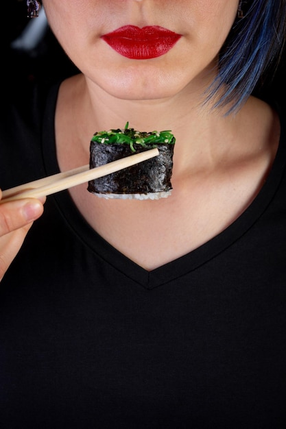 Foto chica de aspecto europeo de piel clara con labios rojos come rollos de sushi sosteniéndolos en sus manos con palos de bambú