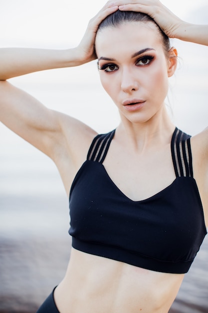 Chica atlética con camiseta sin mangas haciendo un estiramiento en la playa