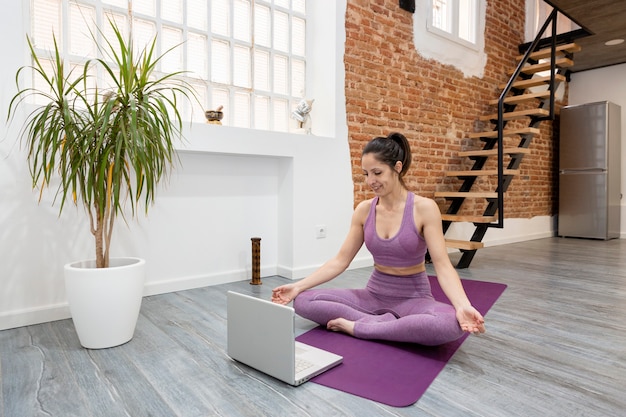 Chica atlética haciendo postura de meditación de yoga frente a su computadora portátil en casa. Concepto de clases online.