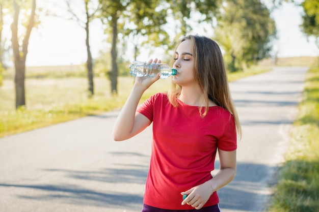 Chica atlética en pantalones cortos violetas y camiseta roja bebiendo agua de una botella de plástico después de hacer ejercicio en la mañana.