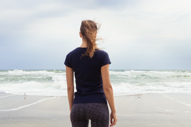 Chica atlética en ropa deportiva de pie en la playa y mirando las olas del mar