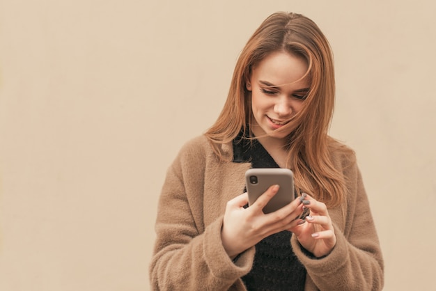 Chica atractiva con un abrigo beige está parado con un teléfono inteligente en las manos de una pared de color beige, mira la pantalla del teléfono inteligente y sonríe