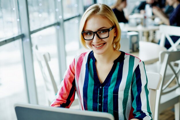 Chica atractiva con cabello claro con camisa colorida y gafas sentado en la cafetería con ordenador portátil, concepto independiente, compras en línea.