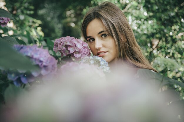 Chica atractiva con cabello largo castaño posando cerca de las flores moradas en el jardín