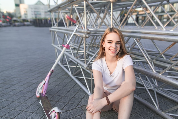 Chica atractiva con una camiseta blanca se sienta en un adoquín con un patinete rosa, mira a la cámara y sonríe.