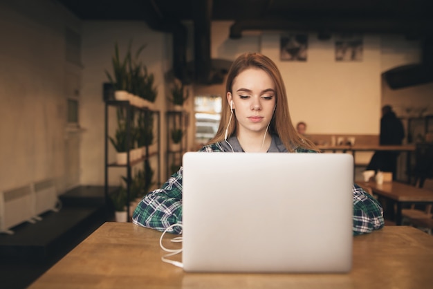 Chica atractiva con una computadora portátil en el café, escucha música en los auriculares y mira la pantalla