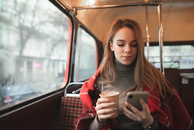 Chica atractiva cubierta con una manta se sienta en un acogedor café con una taza de café en sus manos y mira la pantalla del teléfono inteligente