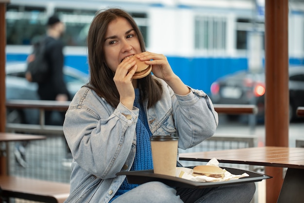 Chica atractiva en estilo casual come una hamburguesa con café sentado en la terraza de verano