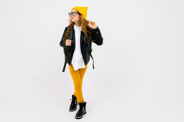 Chica atractiva europea vestida con un sombrero amarillo, gafas y una chaqueta de cuero con una mochila en la espalda en pleno crecimiento en la pared blanca con copyspace