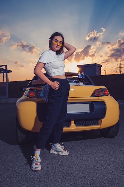 Foto chica atractiva con gafas de sol, pantalones negros superiores y zapatillas de deporte está posando al aire libre cerca del coche descapotable amarillo con dos botellas de refresco en su maletero