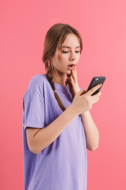 Chica atractiva joven con dos trenzas en camiseta lila sorprendida usando el teléfono celular mientras pasa tiempo sobre fondo rosa