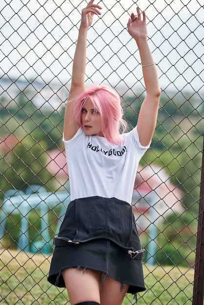 Chica atractiva joven fitness apoyado en la valla, de pie al aire libre. Cabello rosado.