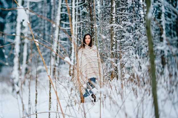 Chica atractiva joven que cubre con una capa caliente posando entre árboles en el bosque de nieve