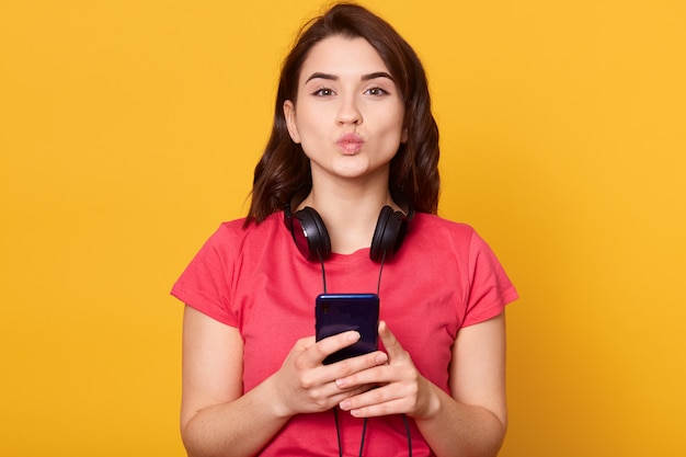 Chica atractiva joven con teléfono en las manos, mostrando un gesto de beso con sus labios, con auriculares alrededor del cuello